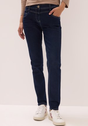 Femme portant un jean en denim bleu foncé et des baskets blanches, se tenant avec une main dans la poche contre un fond beige.