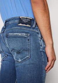 Primer plano de una persona que lleva jeans desgastados de color azul con una etiqueta de "Broken Edge Replay" sobre el bolsillo trasero, con la mano descansando al lado de la pierna.