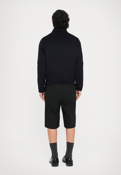 Fekete dzseki magas gallérral, texturált anyagból, rövidített kialakítással, fekete térdig érő nadrággal és sötét zoknikkal párosítva.