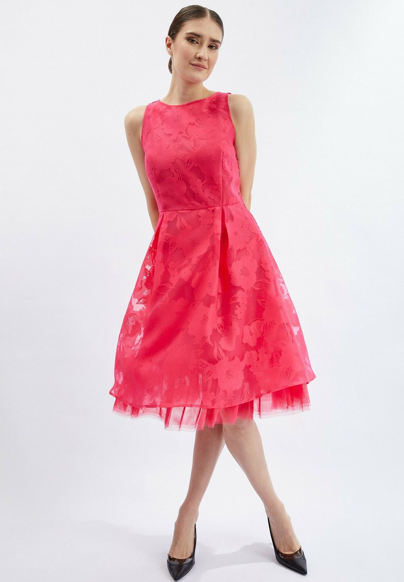 Pinkes ärmelloses Kleid mit floralem Muster, geschichteter Rock und transparenter Unterschicht. Abgerundet mit schwarzen High Heels für den Kontrast.