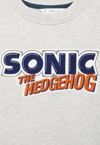 Siva svetrovka z velikim vezenim napisom. Črke so napisane "SONIC" v modri barvi in "THE HEDGEHOG" v oranžni barvi.