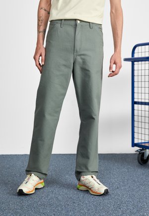 Mand iført lysegrønne bukser med lige ben og hvide sneakers med orange og grønne detaljer, der står på blåt tæppe ved siden af en blå metalkurv.
