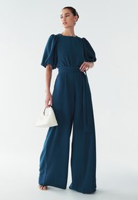 Turquoise jumpsuit met pofmouwen, wijde pijpen en een gestrikte taille. Gecombineerd met een witte driehoekige handtas met een ronde handgreep.