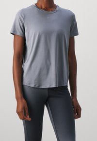 Persona vistiendo una camiseta atlética de manga corta gris claro y mallas ajustadas gris oscuro contra un fondo neutral.