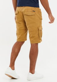 Kahki Cargo-Shorts aus strukturiertem Stoff mit mehreren Seitentaschen und einem Etikett auf der Rückseite, kombiniert mit weißen Sneakers.