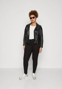 Svart skinn bikerjakke med glidelåser, hvit T-skjorte, svarte tettsittende bukser og sølvfargede sneakers. Modell som har på seg solbriller og står oppreist.