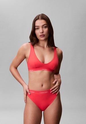 Femme aux cheveux longs bruns portant un soutien-gorge rouge Calvin Klein et des sous-vêtements assortis, debout devant un fond gris uni.