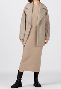 Beige ullblandad kappa med snedskurna slag, dubbelknapplåsning, tillsammans med en ribbad beige stickad klänning och svarta knähöga stövlar.