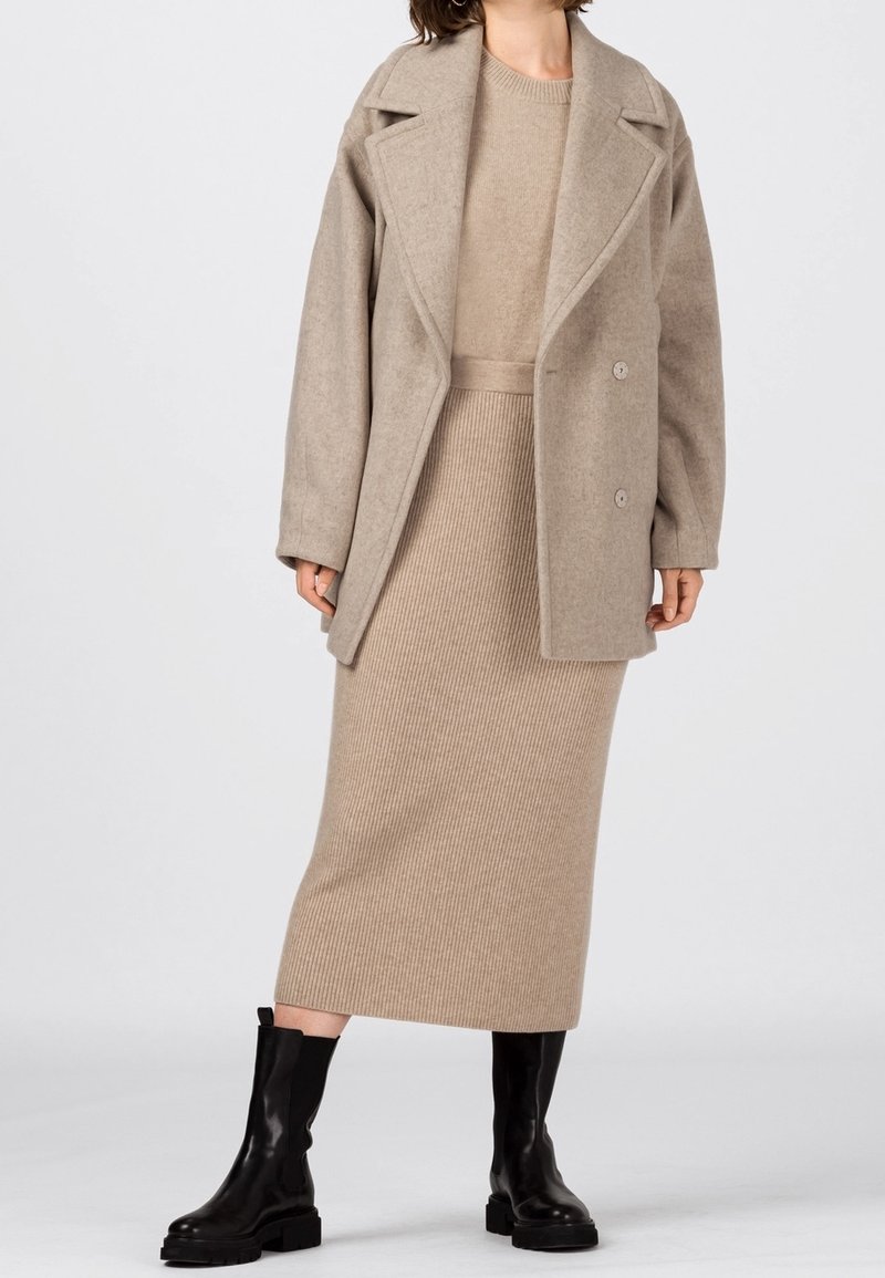 Beige ullblandad kappa med snedskurna slag, dubbelknapplåsning, tillsammans med en ribbad beige stickad klänning och svarta knähöga stövlar.