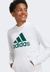 Sudadera blanca con bolsillo canguro, que presenta un gran logo verde de adidas en el pecho. Tejido suave, puños acanalados y capucha.