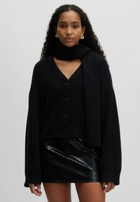 Zwarte gebreide cardigan met V-hals, knopen aan de voorkant en lange mouwen, gecombineerd met een zwarte glanzende leren minirok. Soepele textuur overal.