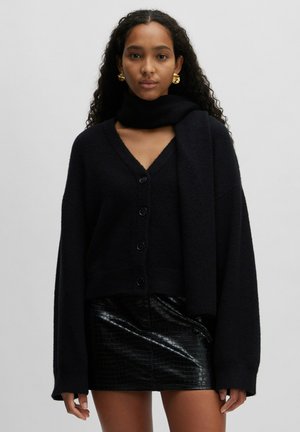 Schwarze gestrickte Strickjacke mit V-Ausschnitt, Knopfleiste und langen Ärmeln, kombiniert mit einem schwarzen glänzenden Leder-Minikleid. Glatte Textur insgesamt.