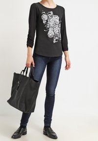 Žena oblečená v černém topu s květinovým vzorem, tmavých skinny džínách, černých mokasínech a držící velkou černou koženou kabelku.