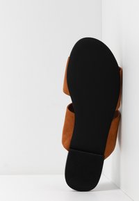 Chaussure plate en daim brun unique, positionnée semelle vers le haut contre un coin blanc, montrant une semelle noire lisse et un talon carré.