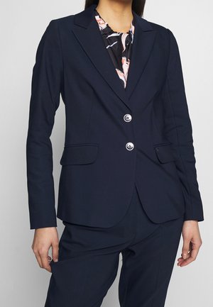 Femme portant un blazer bleu marine ajusté à deux boutons et poches à rabat, sur un chemisier noir à motif floral rose.