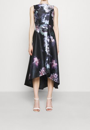 Mouwloze zwarte jurk met paarse en witte bloemenprint, asymmetrische zoom, gedragen met witte enkellaarzen met enkelbandje op effen achtergrond.