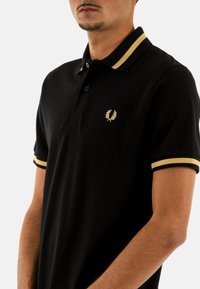 Polo nero con accenti dorati sul colletto e sulle maniche, caratterizzato da un piccolo logo ricamato in oro sul petto, maniche corte e una vestibilità classica.