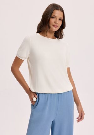 Kobieta ubrana w prostą białą bluzkę z krótkim rękawem i luźne jasnoniebieskie spodnie, pozująca z jedną ręką w kieszeni na neutralnym tle.