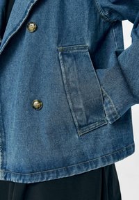 Veste en denim bleue avec un design croisé, boutons en métal et poche latérale. Présente des coutures contrastées et une finition en tissu texturé.