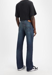 Blå denimjeans med rak bendesign, med en brun läderlapp på baksidan och två bakfickor. Svart t-shirt och sneakers.