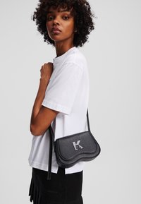 Bolsa transversal de couro preto com design curvo, apresentando um destaque de logotipo proeminente. Usada sobre uma t-shirt branca, mostrando um visual minimalista.