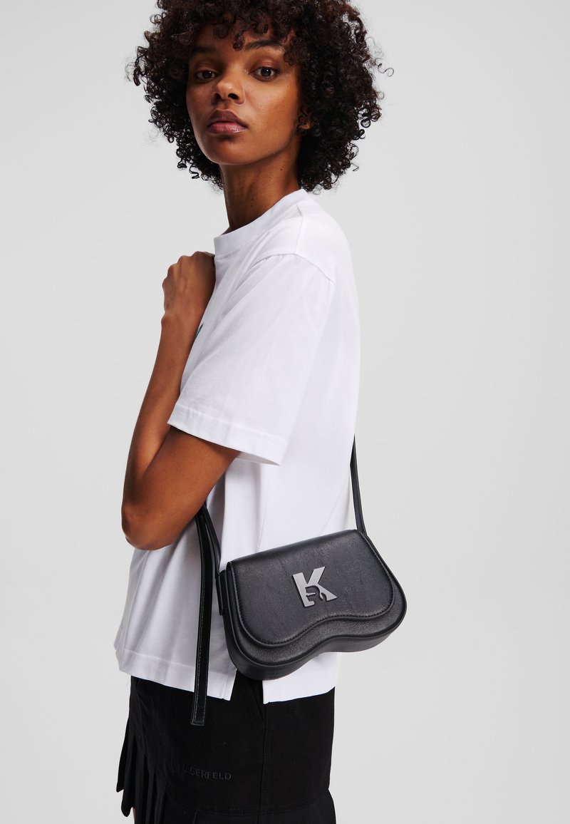 Bolsa transversal de couro preto com design curvo, apresentando um destaque de logotipo proeminente. Usada sobre uma t-shirt branca, mostrando um visual minimalista.