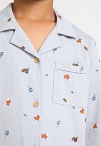 Helles blaues Kurzarmhemd mit Kragen. Verfügt über florale und tierische Druckmuster, Holzknöpfe und eine Brusttasche. Weicher Stoff.