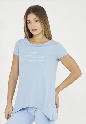 Jeune femme aux cheveux lisses portant un T-shirt bleu clair à ourlet asymétrique avec un message positif et des leggings assortis, debout, main sur la hanche.
