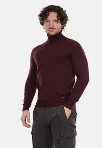 Maglione a collo alto bordeaux realizzato in un morbido tessuto a maglia. Presenta maniche lunghe e una silhouette aderente. Abbinato a pantaloni cargo scuri.