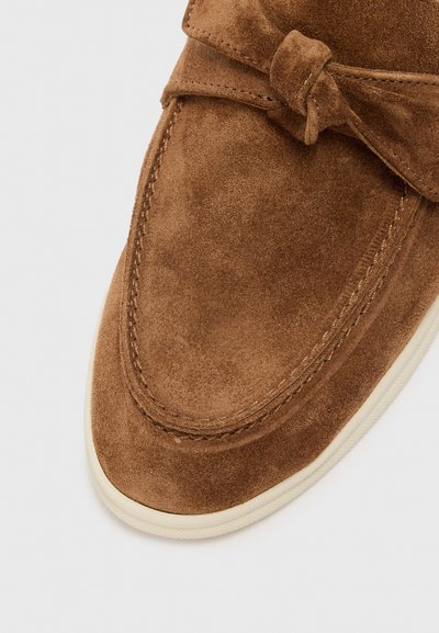 Smeđe antilop mokasinke s ukrasnim čvorom, okruglim vrhom i potplatom krem boje. Glatka tekstura i vidljivo šivanje duž rubova.