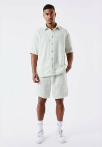 Grünes gemustertes Kurzarmhemd und passende Shorts aus leichtem Stoff. Verfügt über Knöpfe und eine lockere Passform. Weiße Sneakers und Socken runden das Outfit ab.