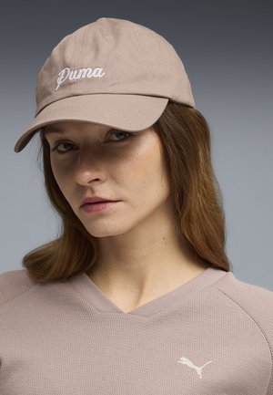 Mujer con cabello castaño claro que lleva una gorra beige de Puma y una blusa texturizada a juego sobre un fondo degradado gris.
