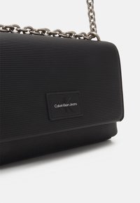 Svart handväska av ribbad material med en silverfärgad kedjerem och en rektangulär logotypmärke med "Calvin Klein Jeans" präglat.