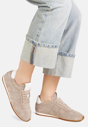 Beżowe zamszowe sneakersy z białymi akcentami, z wiązanym zapięciem, dziurkowanymi detalami i brązową gumową podeszwą, w połączeniu z krótkimi dżinsowymi spodniami.