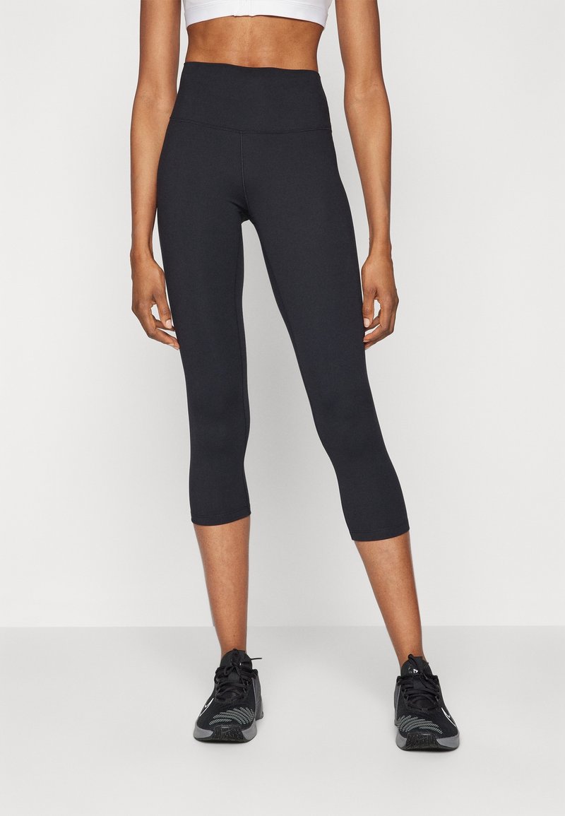 Leggings capri negros con cintura alta, confeccionados con material elástico, que presentan una textura suave y un diseño clásico. Combinados con zapatillas deportivas negras.