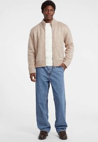 Beige strikket cardigan med høj krave og ribbede manchetter, kombineret med en hvid skjorte og løse blå jeans, afsluttet med mørkebrune sko.