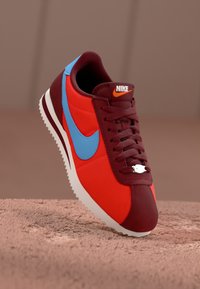 Piros és bordó Nike sportcipők kék részletekkel, texturált szintetikus felsőrésszel, fehér talppal és középső fűzési rendszerrel; Nike logó a nyelven.