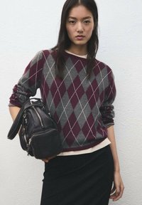 Pulover cu model argyle în nuanțe de maro închis și gri, cu manșete striate, asortat cu o geantă neagră cu fermoar. Modelul poartă o fustă neagră dedesubt.