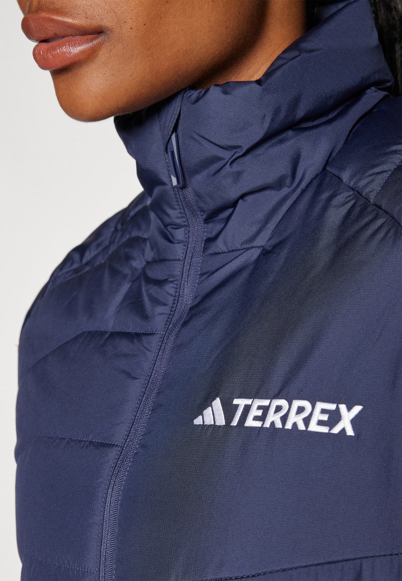 Doudoune bleu marine avec col haut, détails zippés, et logo "TERREX" blanc sur la poitrine, avec une finition en tissu lisse et texturé.