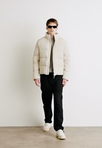 Geacă puffer bej deschis cu un design scurtat, textură matlasată, guler în picioare, asortată cu pantaloni negri și adidași albi.