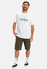 Bílý bavlněný tričko s modrým logem "Quiksilver" uprostřed, doplněné olivově zelenými cargo kraťasy. Černé tenisky a bílé nízké ponožky dotvářejí celkový vzhled.