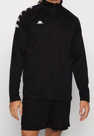 Mężczyzna ubrany w czarną sportową bluzę z długim rękawem z białymi logotypami Kappa na klatce piersiowej i rękawach, zestawioną z czarnymi szortami, stojący na jednolitym tle.