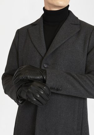 Abrigo de lana gris con un solo botón, llevado sobre un cuello de tortuga negro. Guantes de cuero negros con detalles de costura cubriendo las manos.
