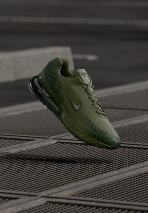 Grøn Nike Air Max sneaker med sort sål og snørebånd, vist flyvende over en metalskæreflade i en udendørs bymæssig setting.