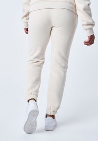 Pantalones de chándal beige con puños elásticos, hechos de tela suave. Llevados con zapatillas deportivas blancas. Diseño sencillo sin patrones ni acentos visibles.