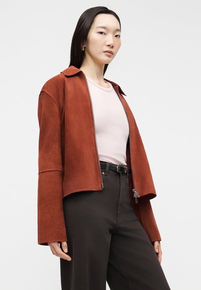 Γυναίκα που φοράει σουέντ μπουφάν σε σκουριά απόχρωση πάνω από ανοιχτό ροζ πουκάμισο και μαύρο παντελόνι, στέκεται μπροστά σε ανοιχτόχρωμο, απλό φόντο.