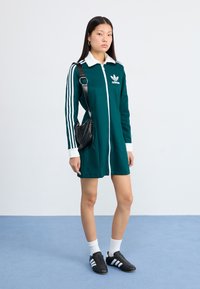 En ung kvinna står på en blå matta, iförd en grön klänning med dragkedja och vita detaljer samt svarta Adidas-skor, och håller en liten svart väska.