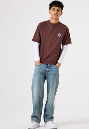 Camiseta marrón de manga corta con un gráfico azul en el pecho, superpuesta a una camisa blanca de manga larga; combinada con jeans azul claro y zapatillas blancas.