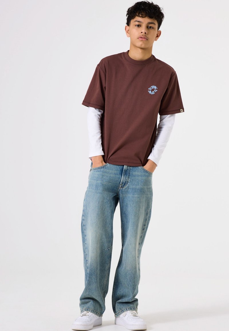 Camiseta marrón de manga corta con un gráfico azul en el pecho, superpuesta a una camisa blanca de manga larga; combinada con jeans azul claro y zapatillas blancas.