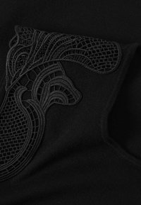 Fekete szövet, amelyen bonyolult fekete csipke részletek találhatók geometrikus mintával és sima textúrával, a ruhadarab egyik sarkában elhelyezve.